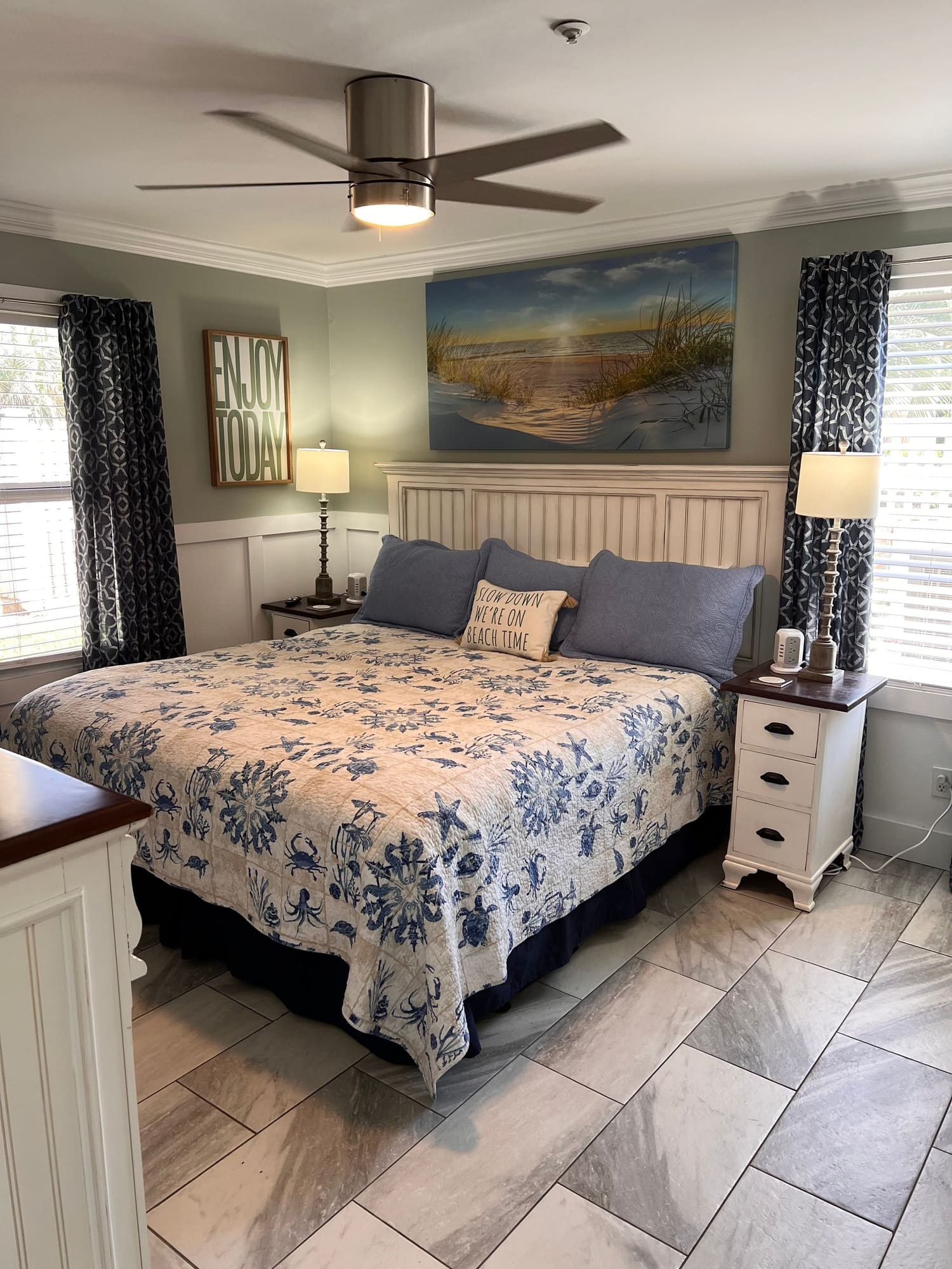 Modern bedroom with blue and cream bedding, ceiling fan, coastal artwork, and patterned curtains